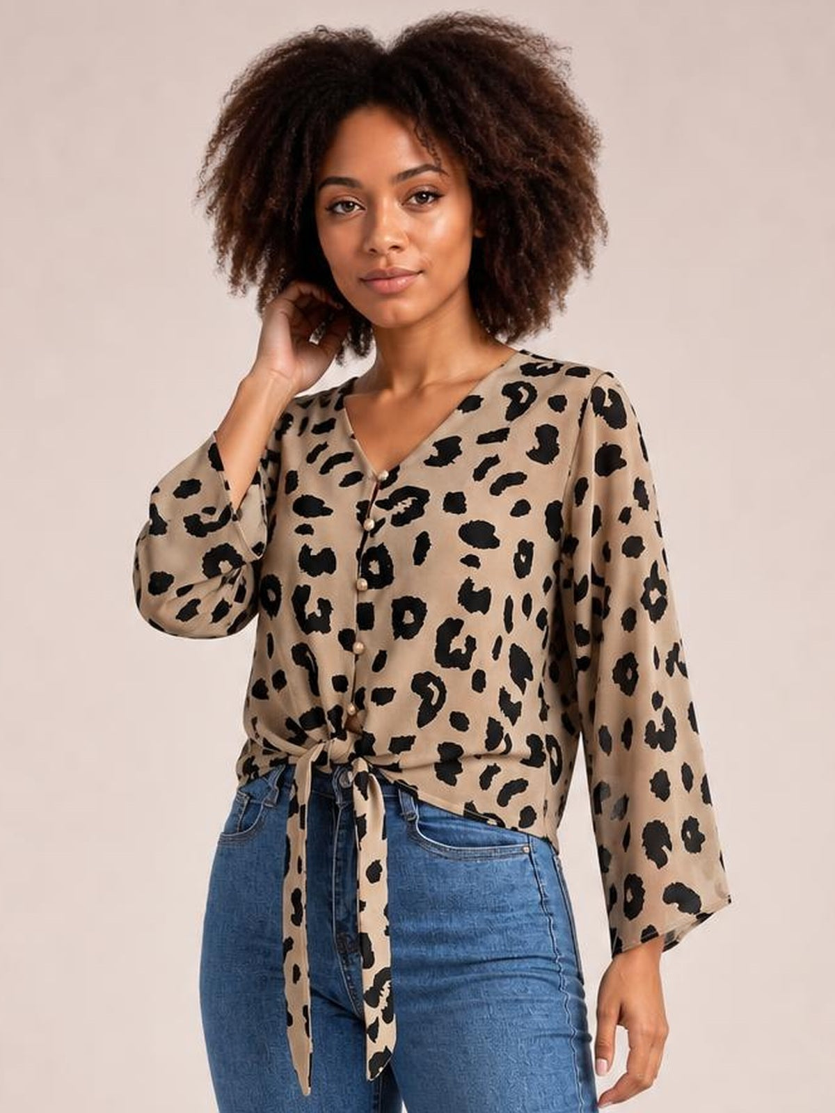 Model wearing a beige leopard-print blouse with a V-neck, button front, long sleeves, and a tied waist, posing with one hand in her hair.