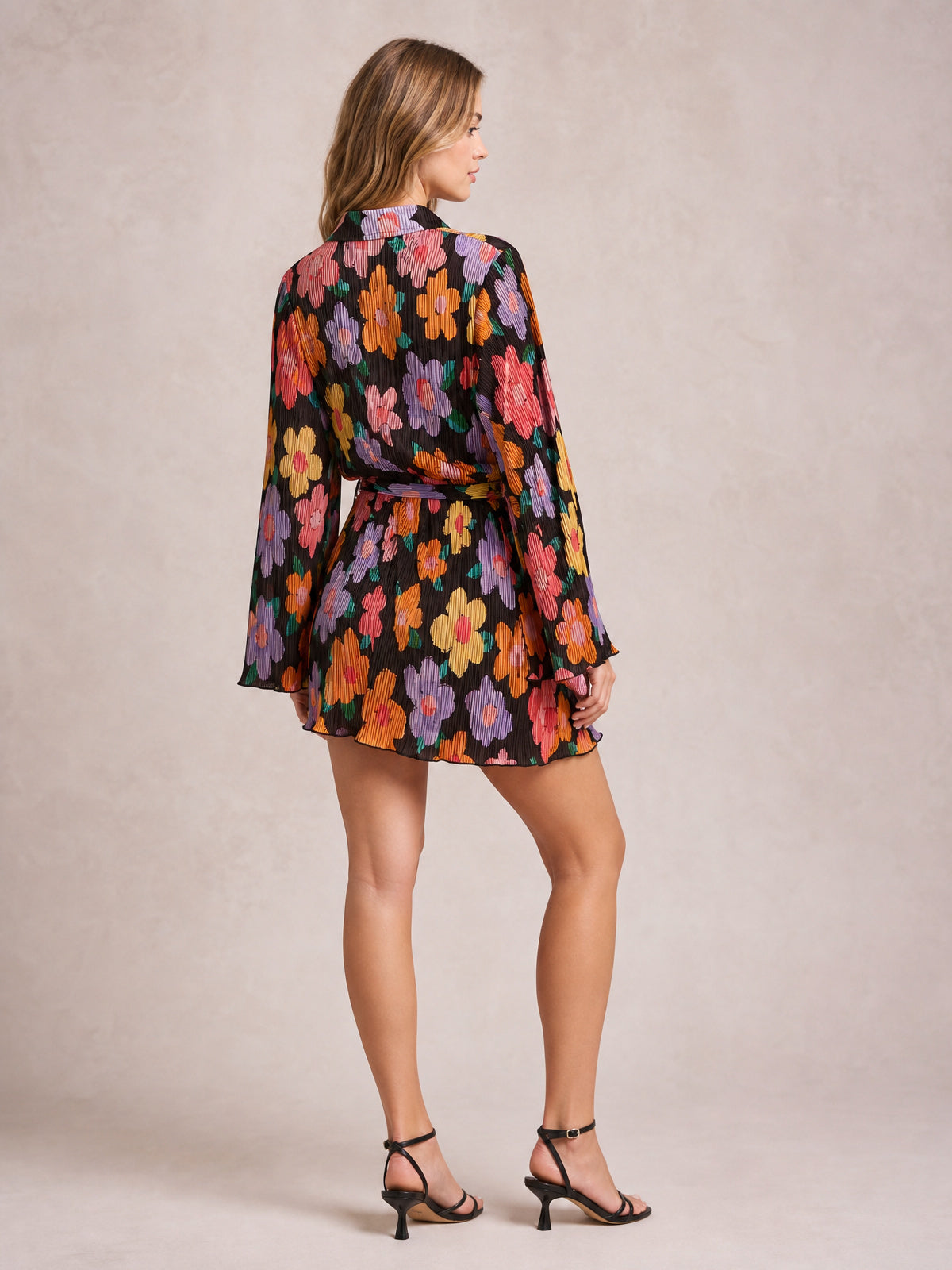 Model poses in a black floral tied button-up dress with long sleeves, a defined waist, softly flared short silhouette, and gathered cuffs.