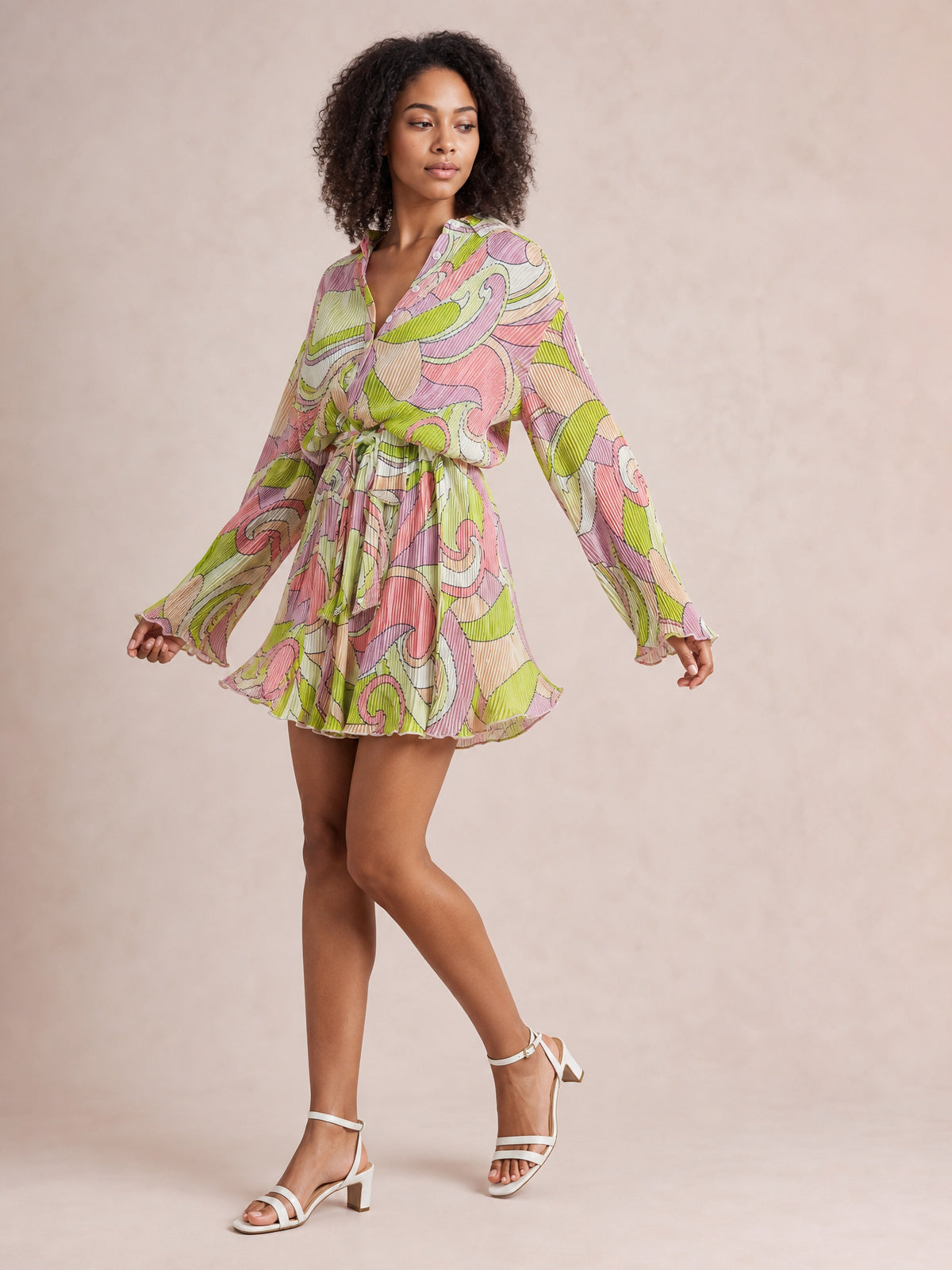 Model poses in a colorful paisley tied button-up dress with a V-neckline, long gathered-cuff sleeves, defined waist, and flared short skirt.