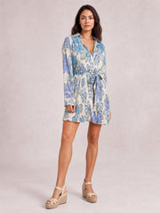 A model stands in a blue and white floral button-up dress with a V-neckline, tied waist, long sleeves, and a breezy short flared silhouette.