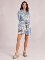 A model stands in a blue and white floral button-up dress with a V-neckline, tied waist, long sleeves, and a breezy short flared silhouette.