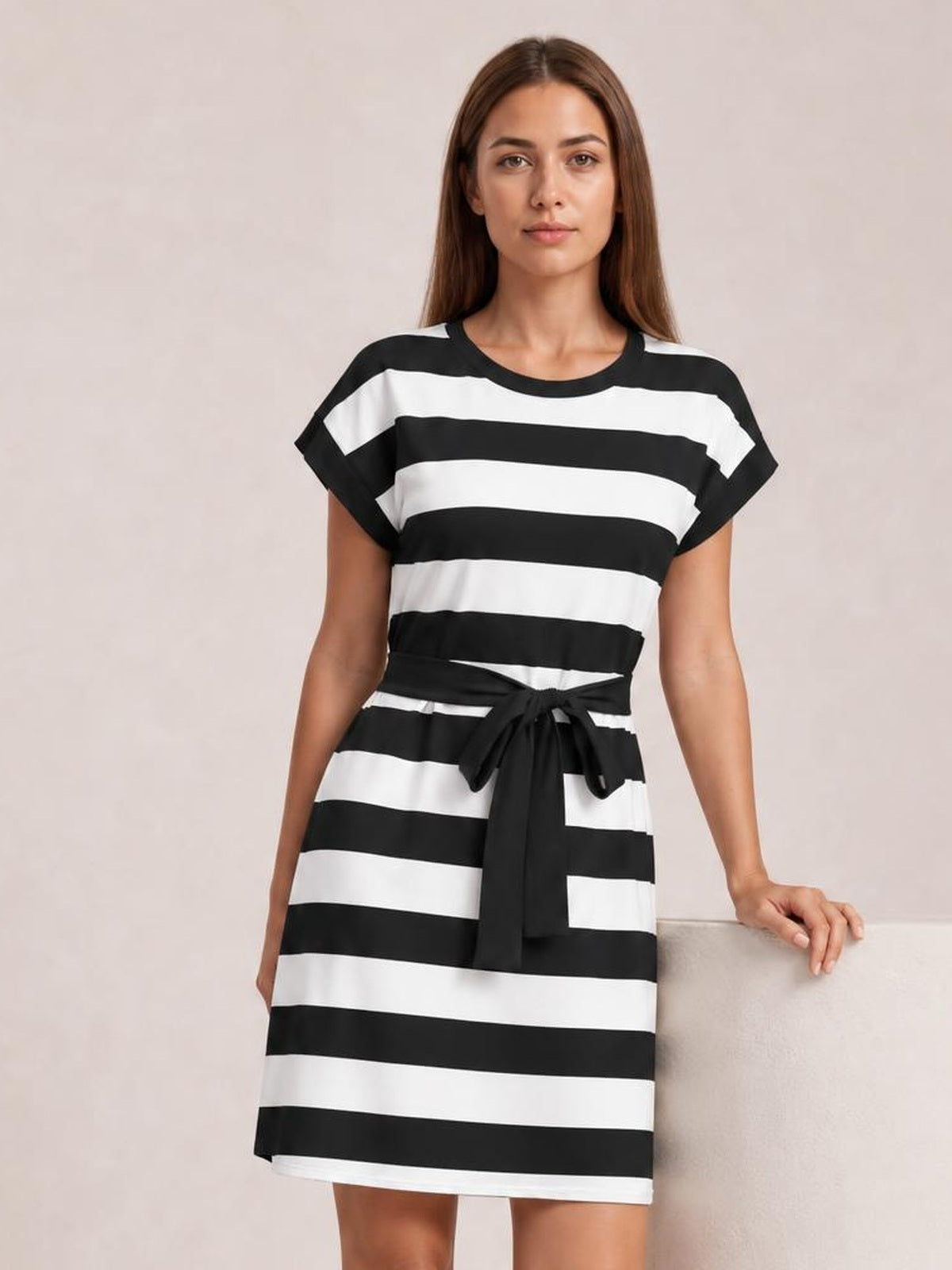 A model stands in a black-and-white contrast striped mini tee dress with short sleeves, a crew neckline, relaxed silhouette, and tied waist.
