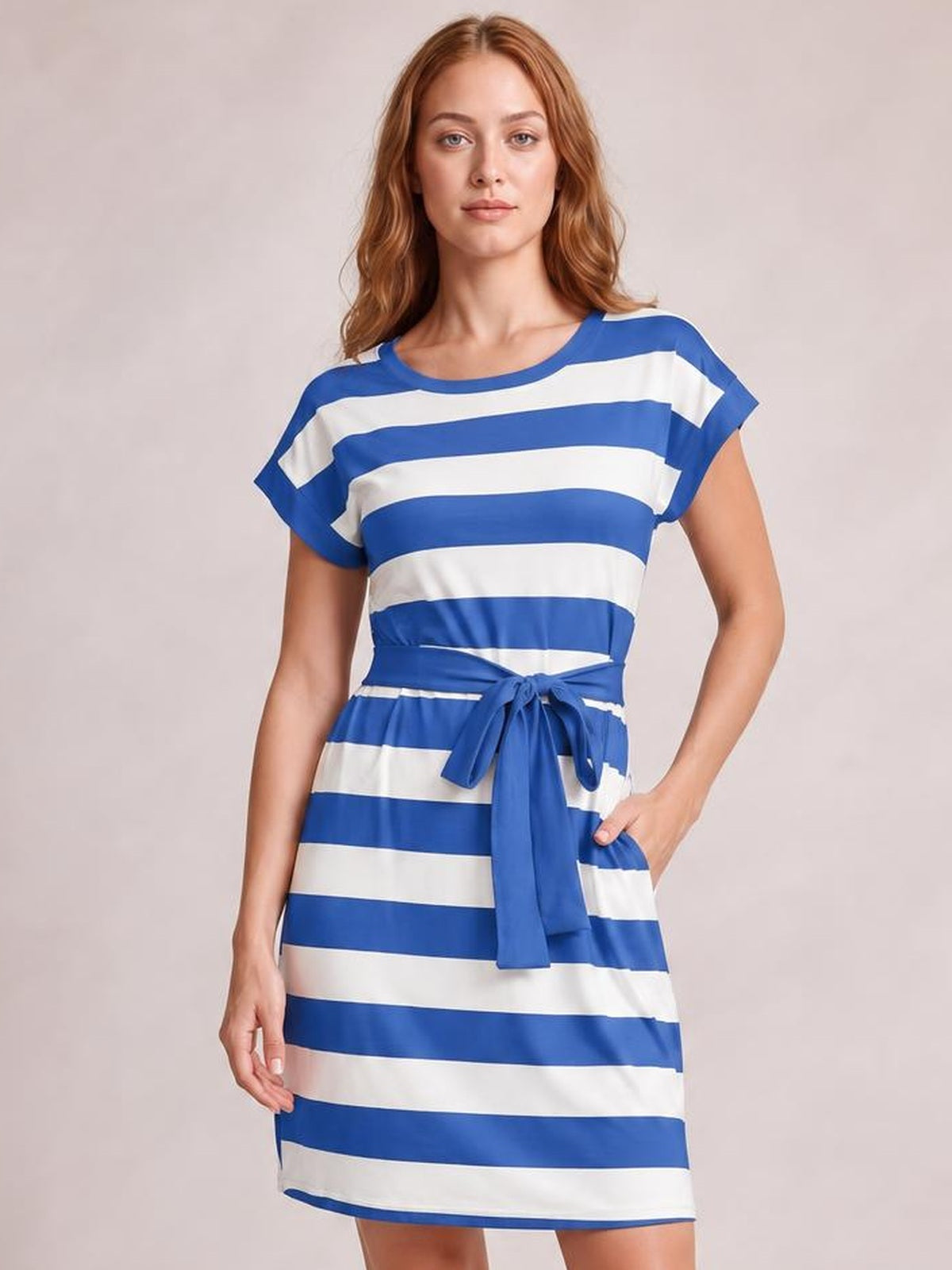 A model wears a blue-and-white contrast striped mini tee dress with short sleeves, a round neckline, relaxed silhouette, and tied waist sash, posing with one hand in a pocket.