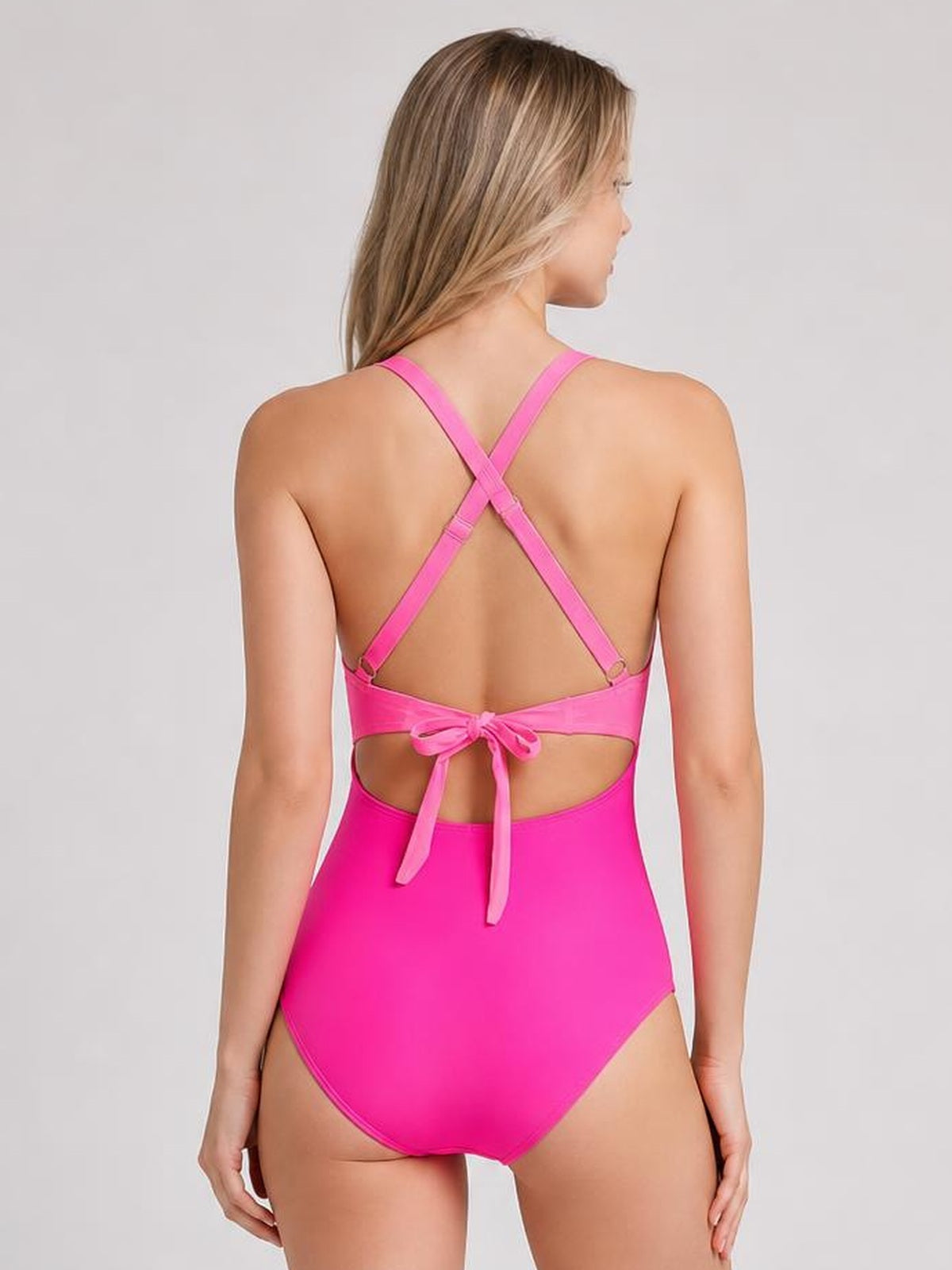 Model shown from the back in a bright pink one-piece swimsuit with crisscross straps, a tied waist, open-back cutout, and a sleek fitted silhouette.