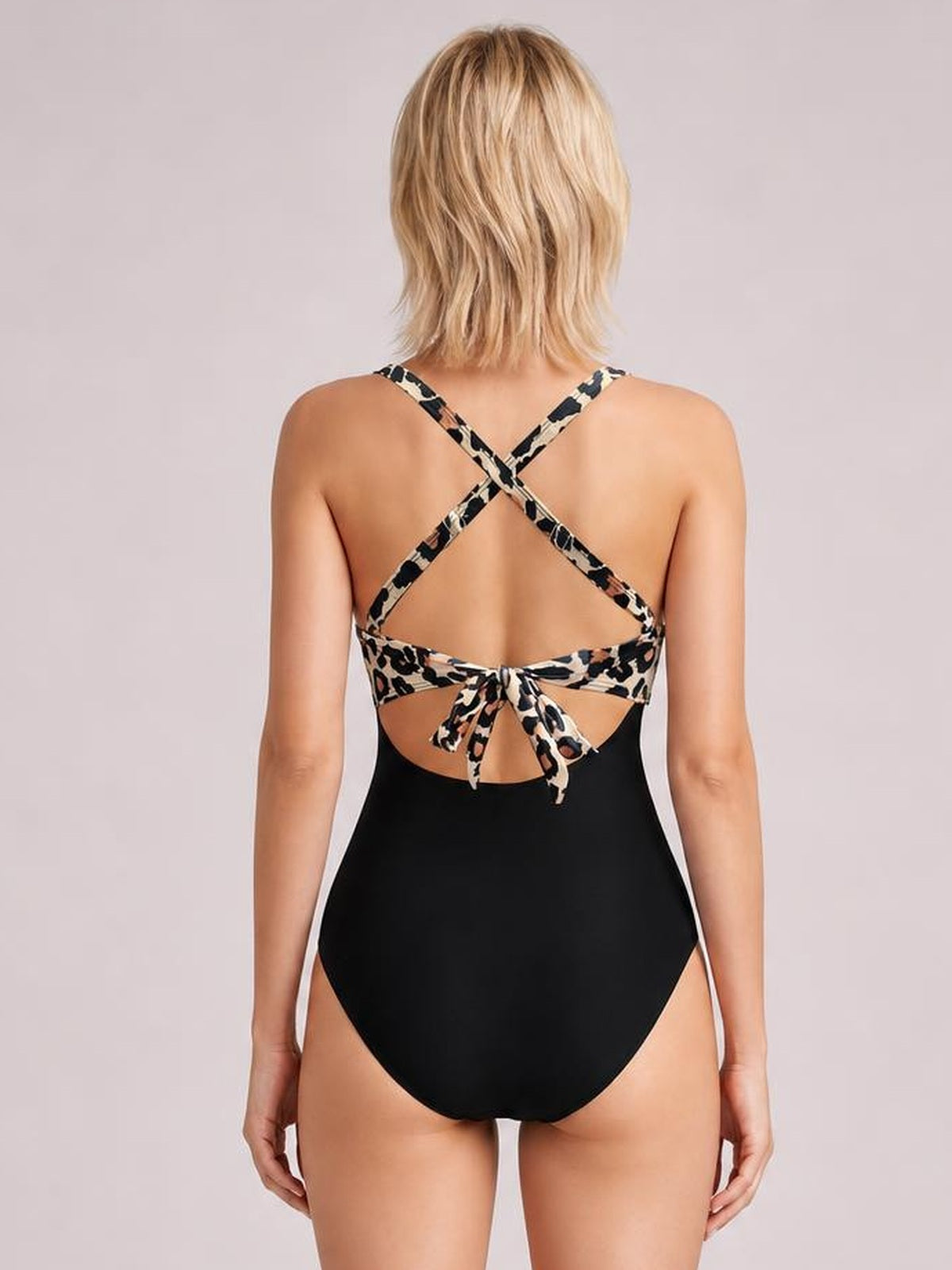 Model shown from the back wearing a black one-piece swimsuit with leopard-print crisscross straps, tied cutout back, and a fitted silhouette.