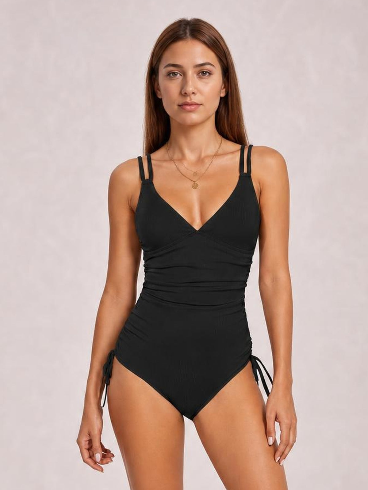 Model stands facing forward in a black tied cutout one-piece swimsuit with a plunge neckline, double shoulder straps, ruched waist, and side ties.