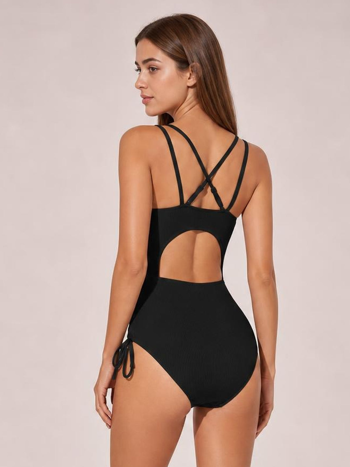 Model shown from behind wearing a black solid one-piece swimsuit with crisscross shoulder straps, a large back cutout, and a tied side detail.