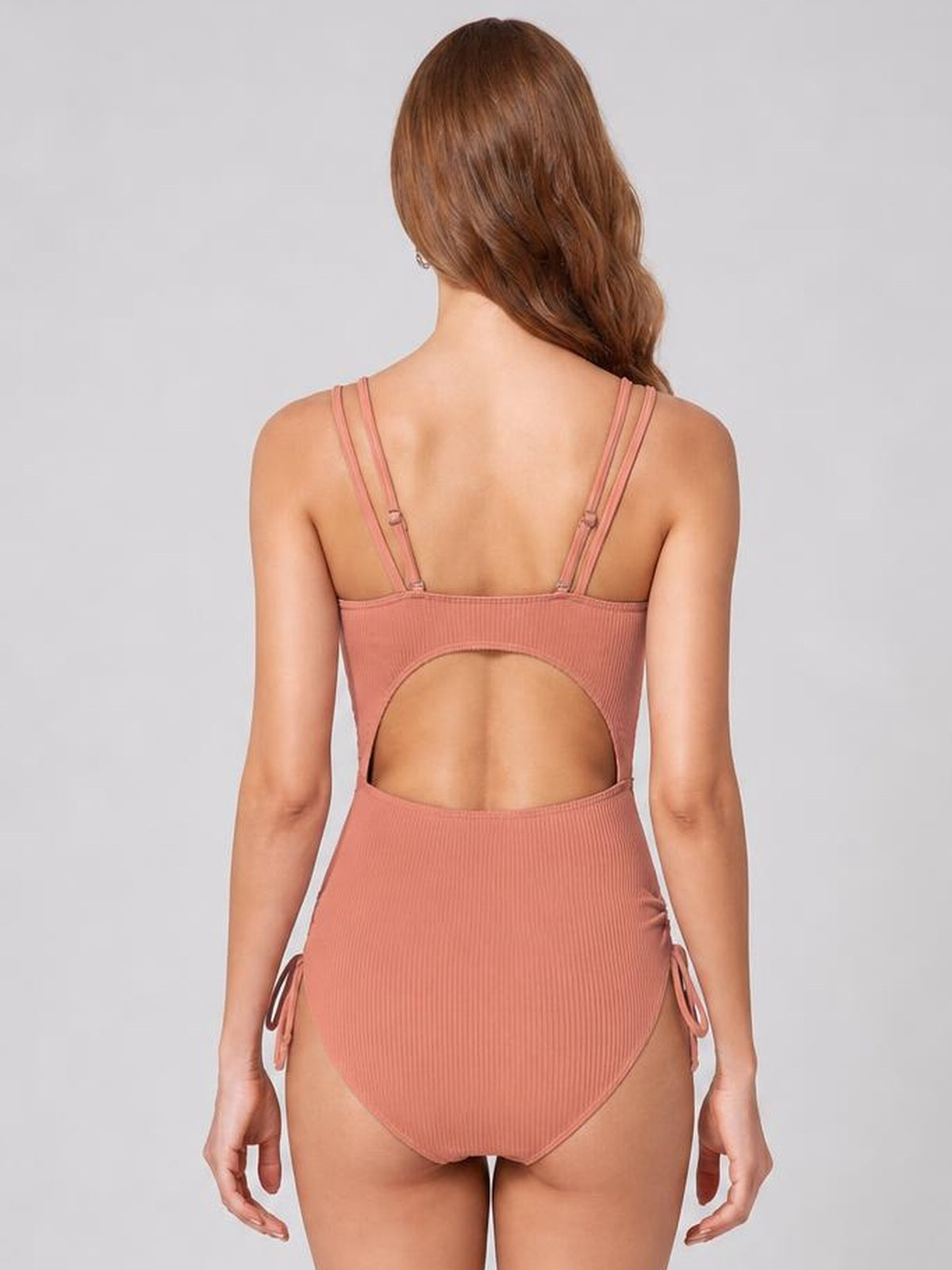 A model poses from behind in a dusty rose tied cutout one-piece swimsuit with double shoulder straps, ribbed fabric, side ties, and a high-cut silhouette.