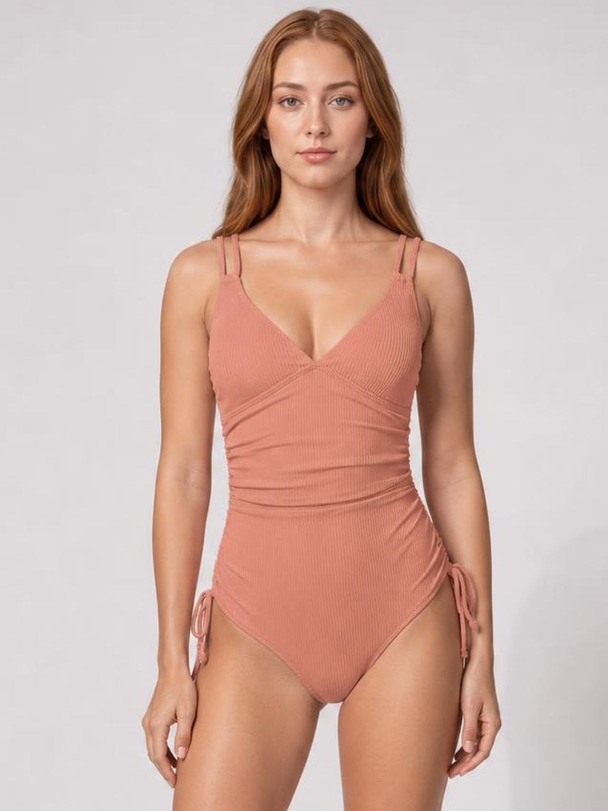 Model stands front-facing in a coral ribbed one-piece swimsuit with a plunge neckline, double shoulder straps, ruched shaping, and tied side details.