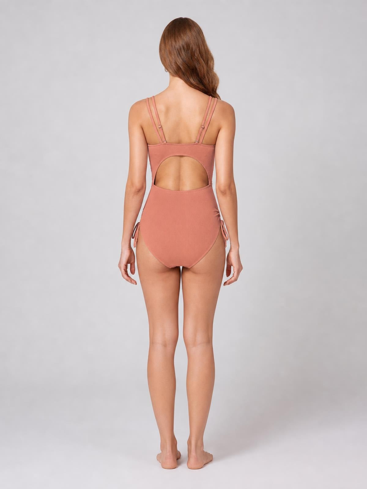 Back view of a model standing in a dusty rose Tied Cutout One-Piece Swimsuit with a large open-back cutout, double straps, ribbed texture, and side ties.