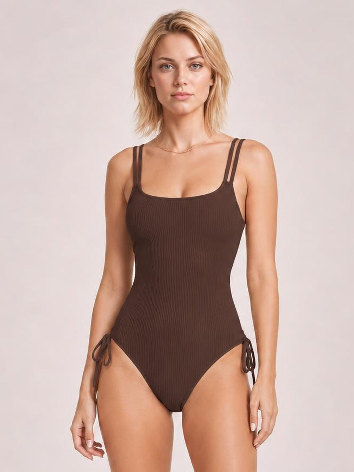 A model stands in a deep brown tied cutout one-piece swimsuit with double straps, a square neckline, ribbed texture, high-cut legs, and side ties.