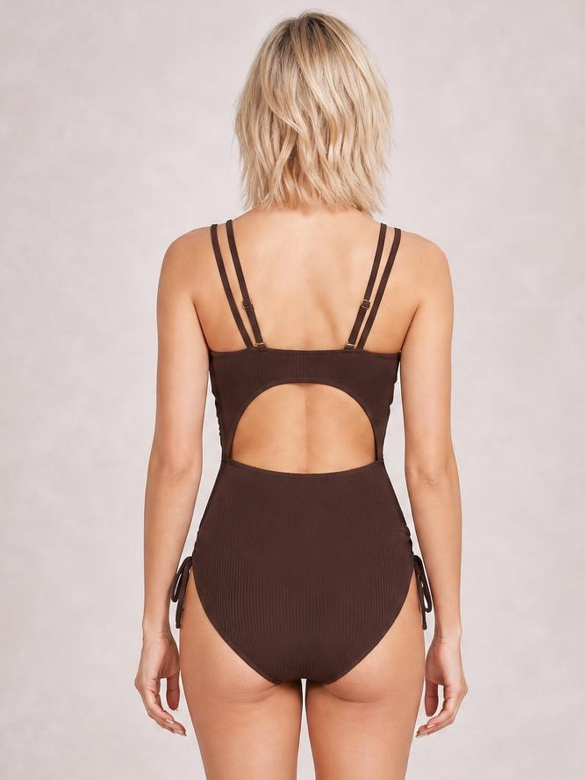 Model stands facing away in a solid brown one-piece swimsuit with a large back cutout, double adjustable straps, ribbed fabric, and tied side details.