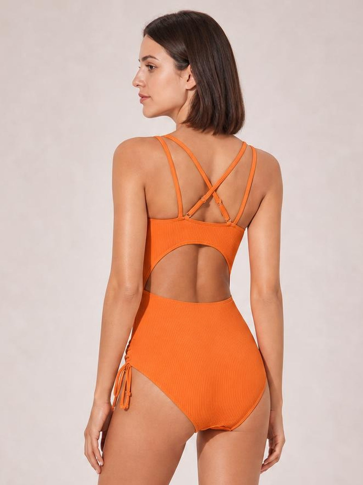 Model shown from the back in a solid orange tied cutout one-piece swimsuit with crisscross straps, high-cut silhouette, and side ties.
