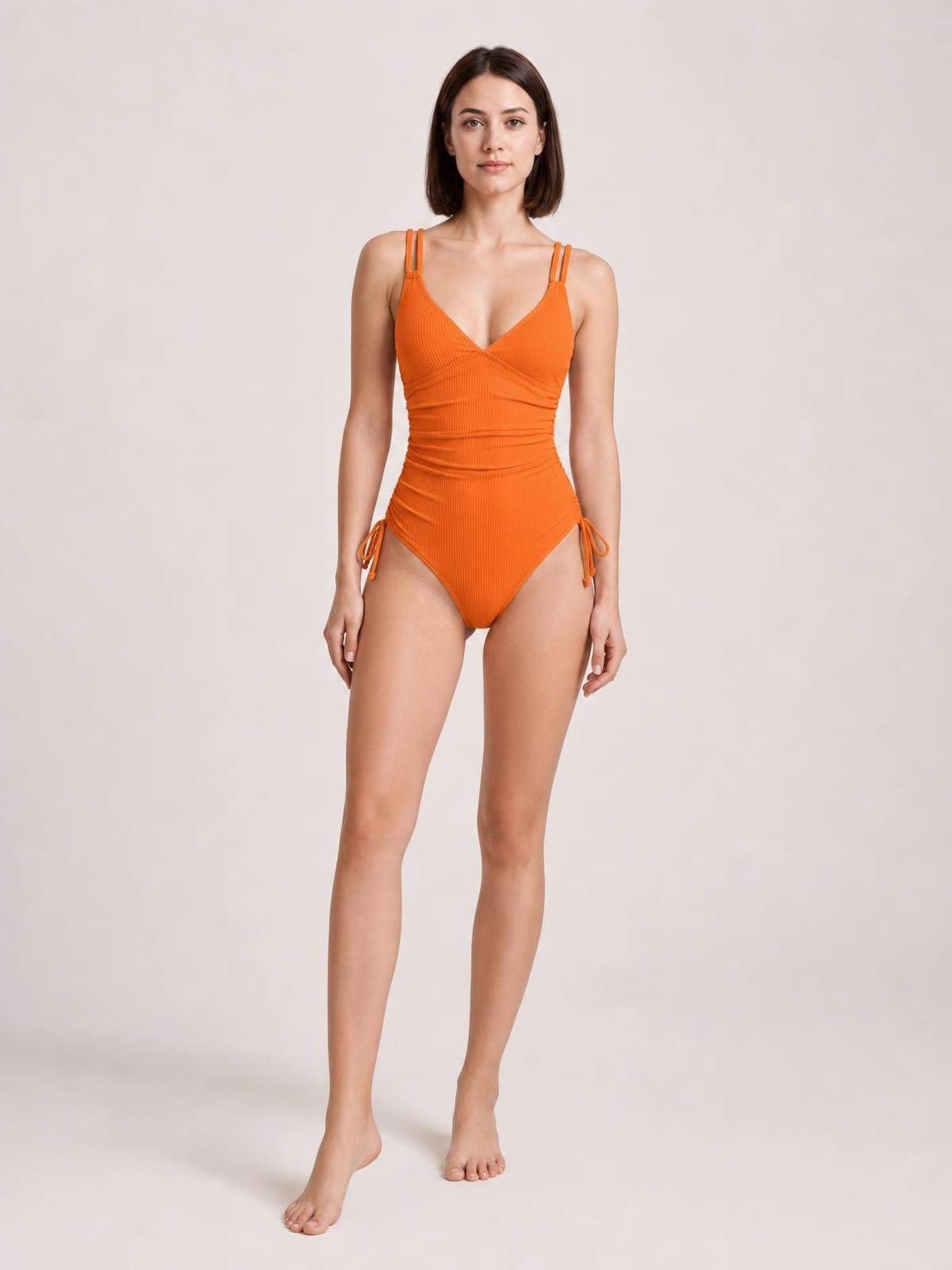 Front-facing model stands in an orange tied cutout one-piece swimsuit with a plunge neckline, double straps, gathered waist, side ties, and high-cut legs.