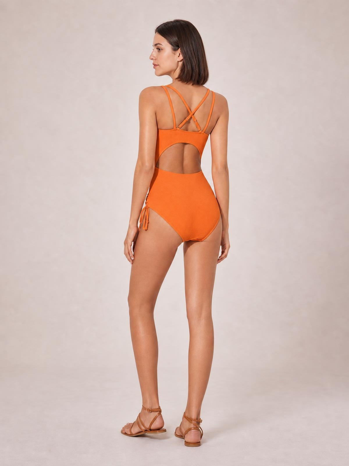 Model stands facing away in an orange tied cutout one-piece swimsuit with crisscross straps, a large back cutout, and a high-cut silhouette.