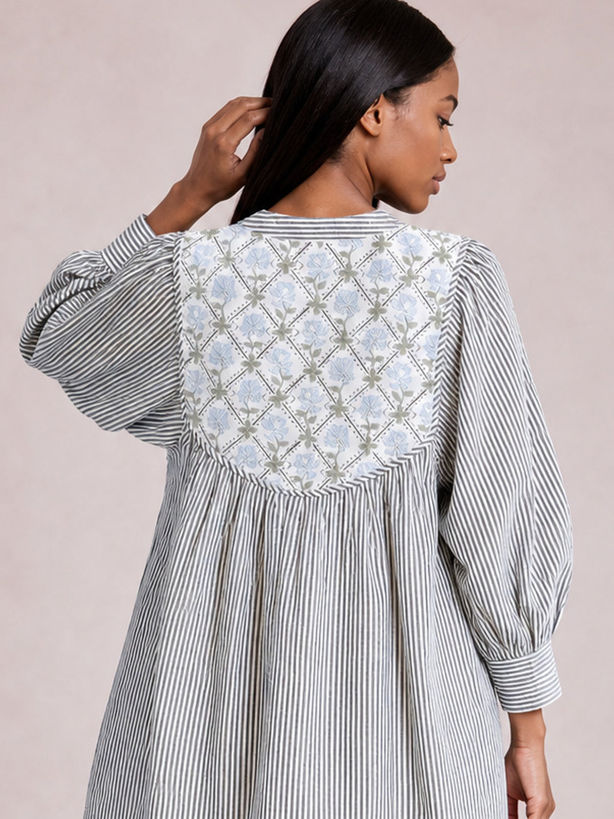 A model poses from the back in a blue-and-white striped dress with a contrasting floral yoke, gathered waist, voluminous long sleeves, and banded cuffs.