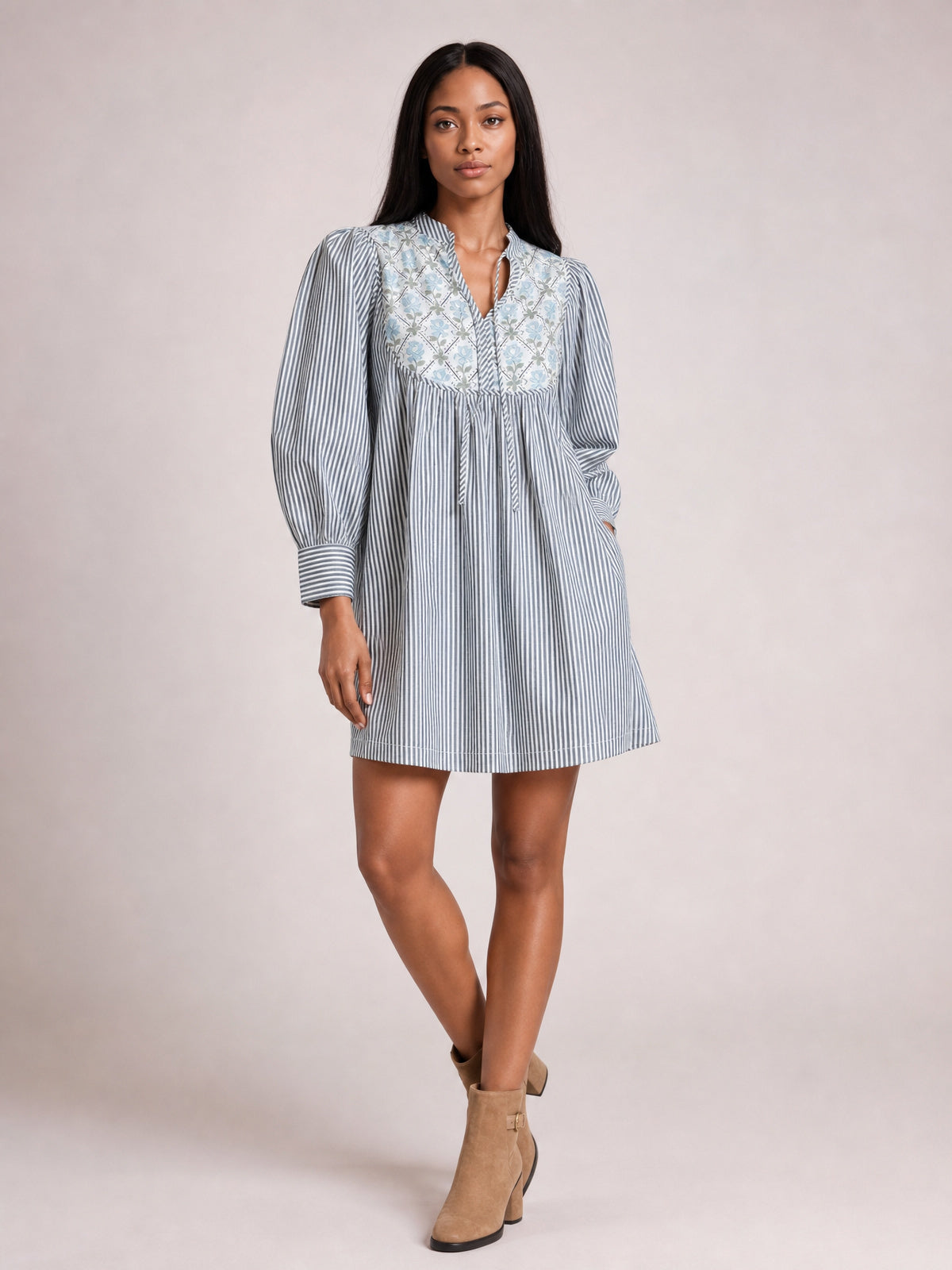 A model poses with crossed legs in a blue-and-white striped floral-yoke dress featuring a V-neck tie, gathered waist, voluminous sleeves, banded cuffs, and an above-knee silhouette.