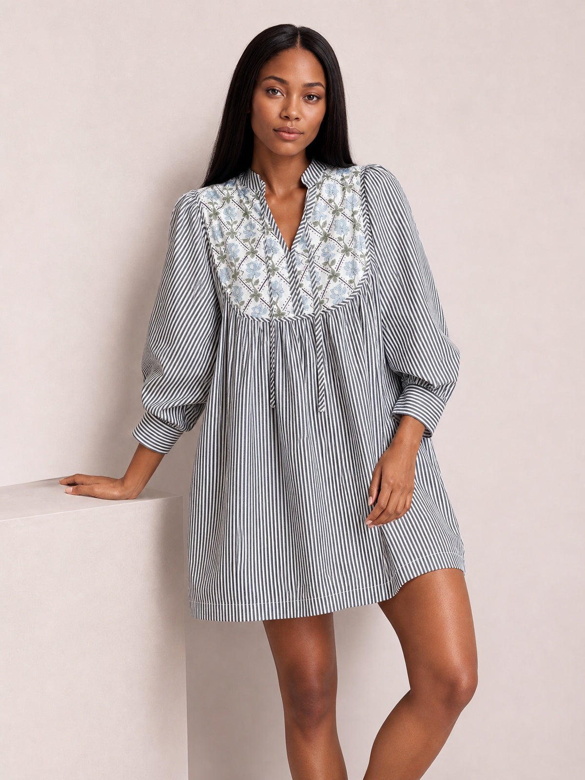 A model leans with one hand resting beside her in a gray-and-white striped floral-printed dress with a V-neck tie, gathered waist, voluminous sleeves, and banded cuffs.