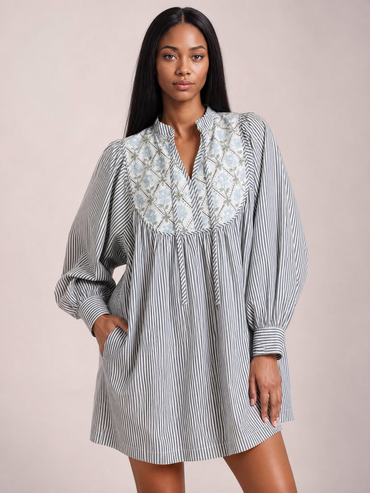 A model stands in a gray-and-white striped dress with a floral yoke, V-neck tie, gathered waist, voluminous long sleeves, banded cuffs, and an above-the-knee silhouette.