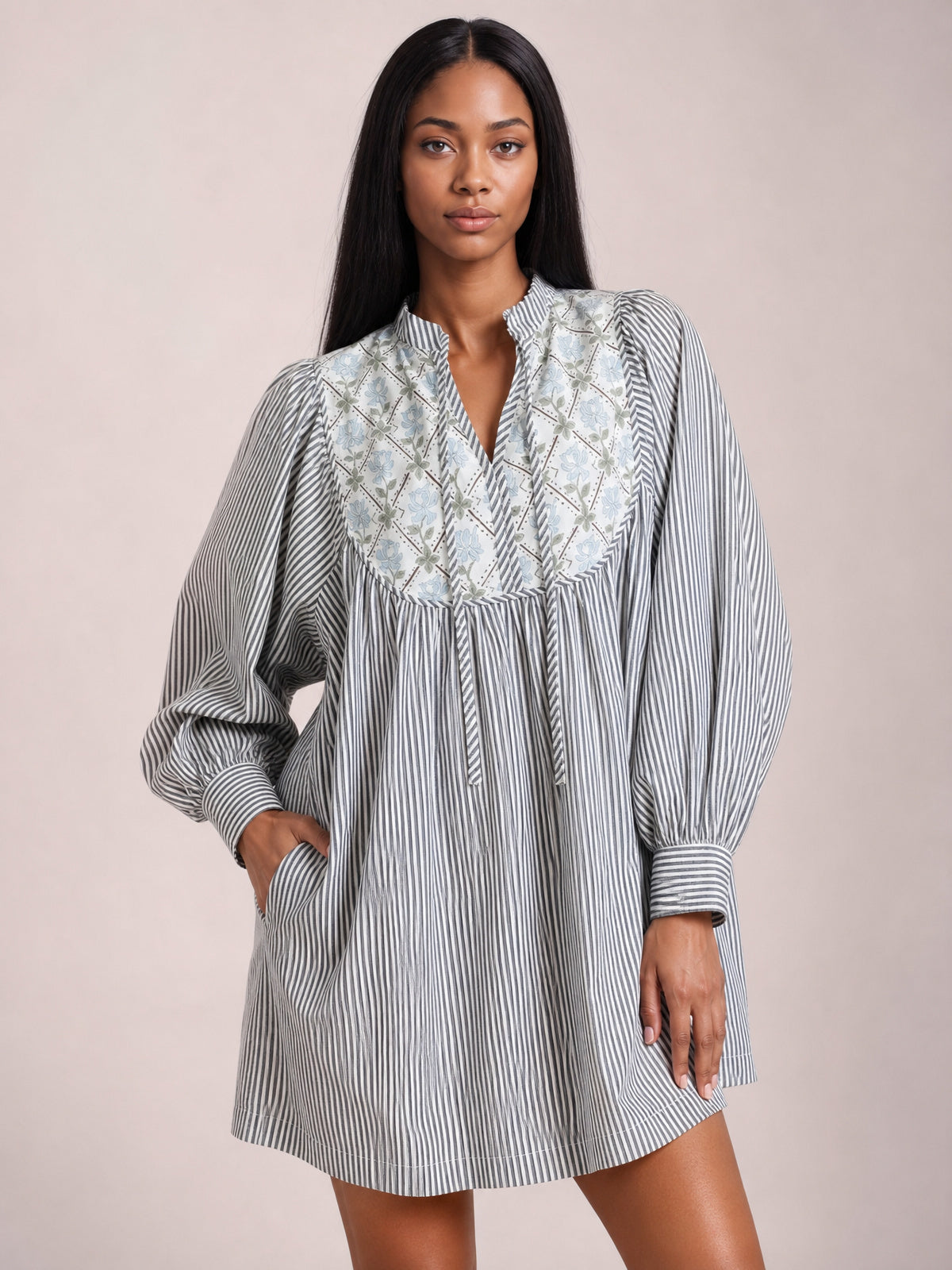 A model stands in a gray-and-white striped dress with a floral yoke, V-neck tie, gathered waist, voluminous long sleeves, banded cuffs, and an above-the-knee silhouette.
