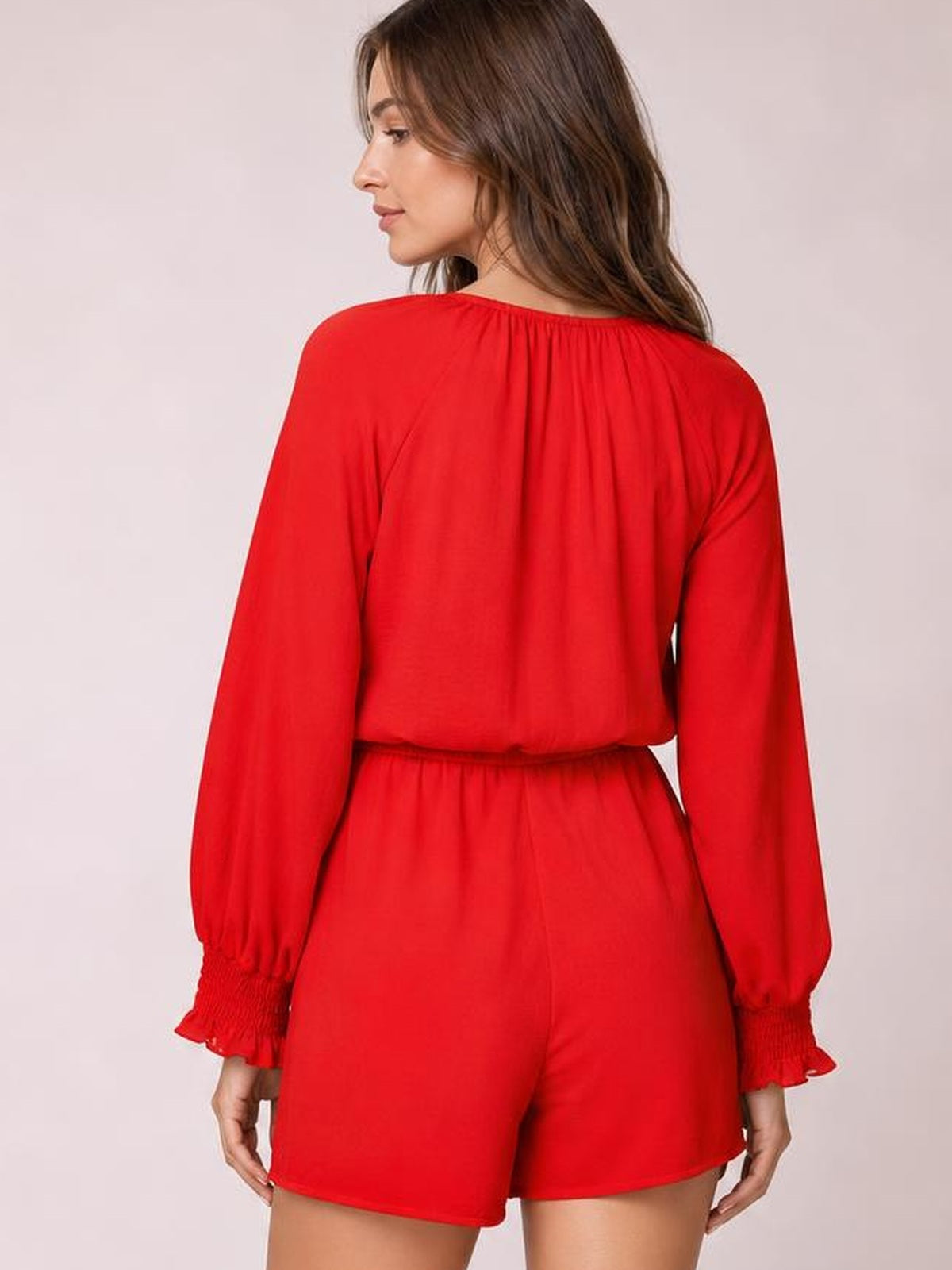 A model stands facing away in a solid red romper with a relaxed silhouette, long lantern sleeves, flounce cuffs, gathered waist, and short legs.