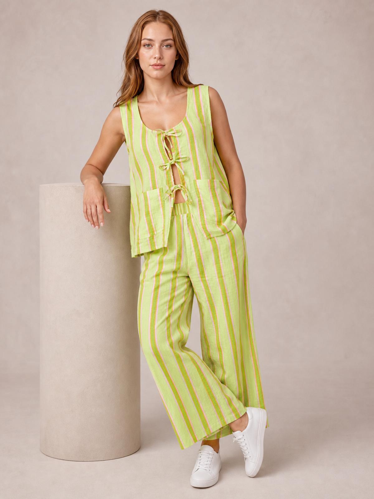 A model poses in a lime green and pink striped top and pants set, featuring a sleeveless tied-front top, wide-leg pants with pockets, and crossed legs.