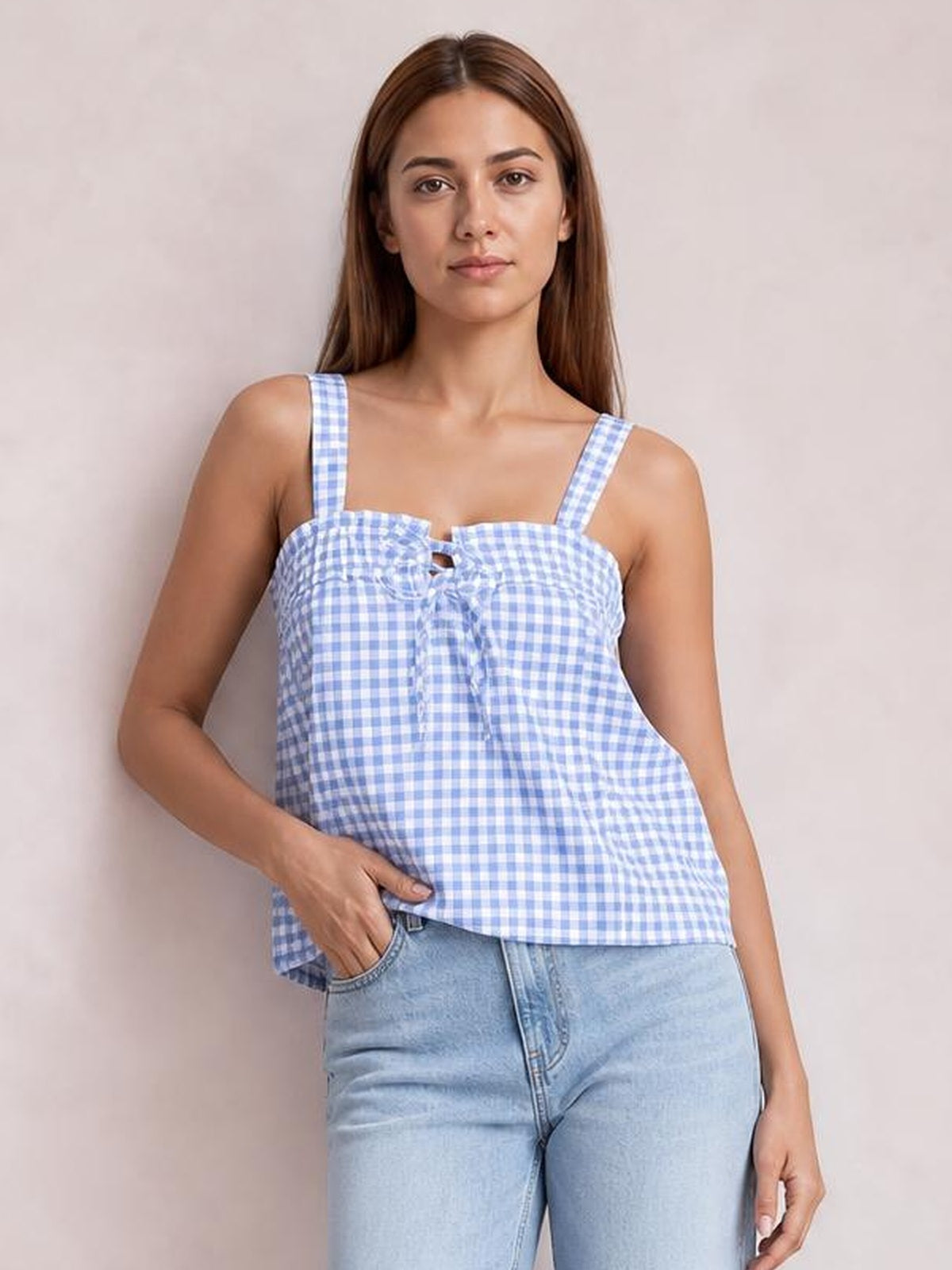 Model poses with one hand in her pocket wearing a blue gingham textured cami with square neckline, shoulder straps, tied front, and a relaxed flared silhouette.