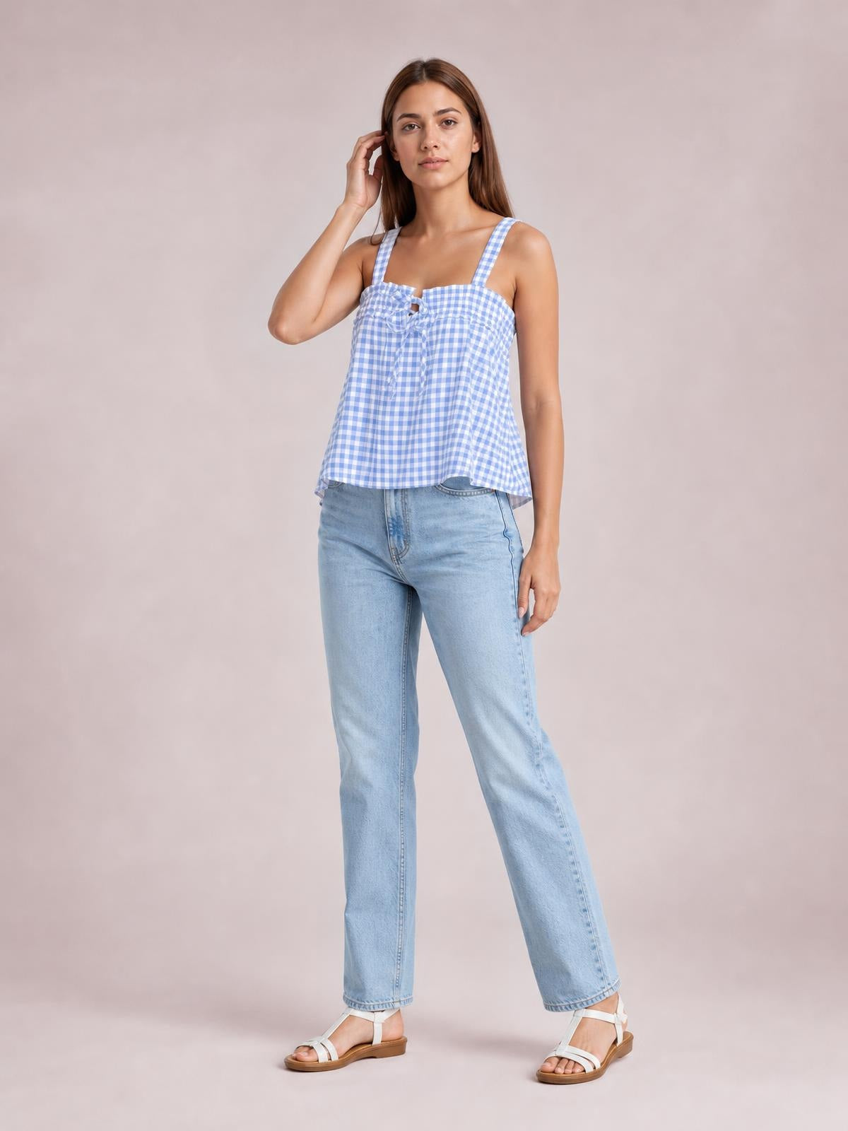 A model poses in a blue-and-white textured gingham cami with a square neckline, shoulder straps, tied front detail, and relaxed flared silhouette.