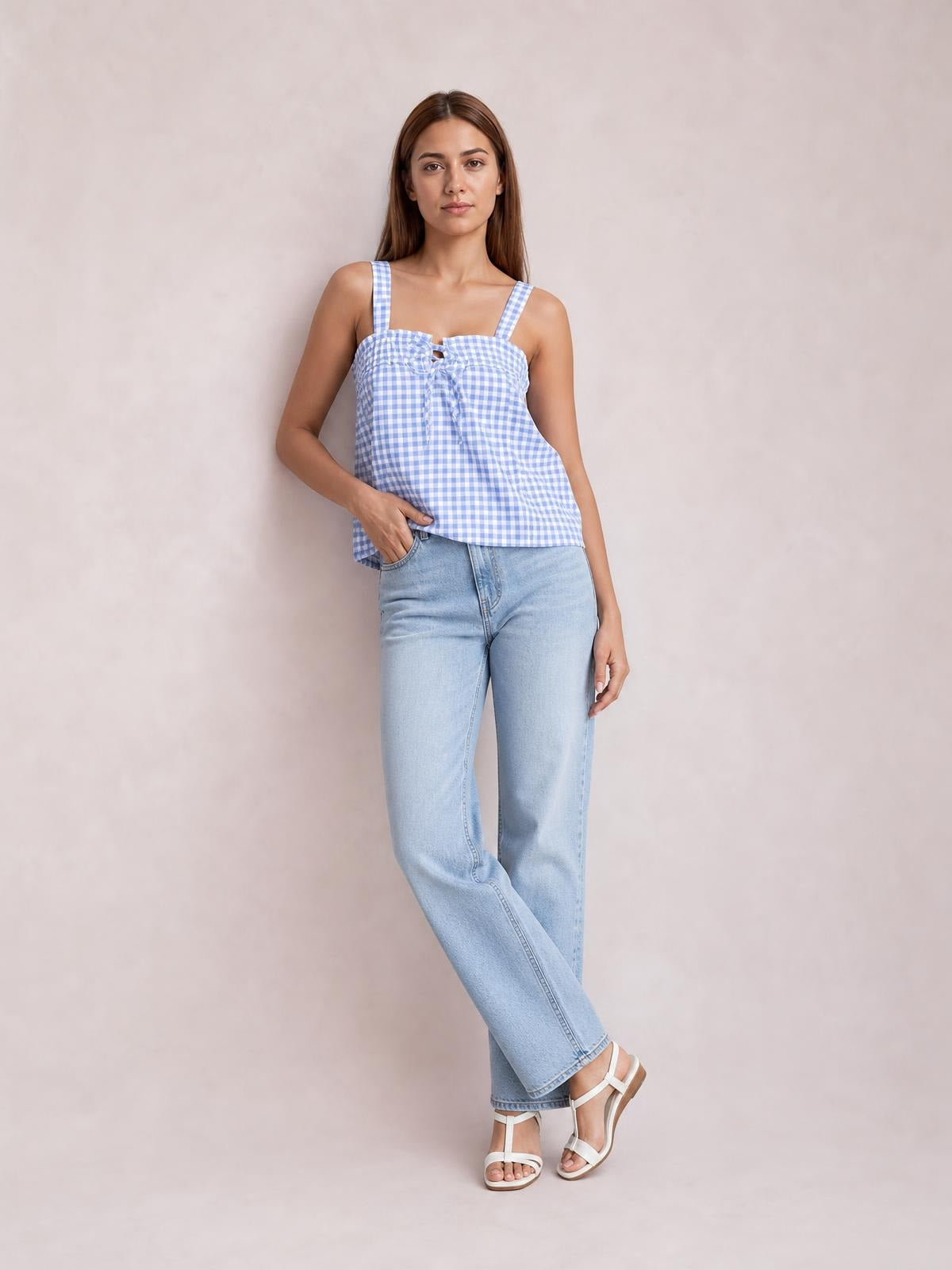 Model wears a light blue and white textured cami with a square neckline, shoulder straps, front tie, and relaxed flared silhouette while standing with crossed legs.
