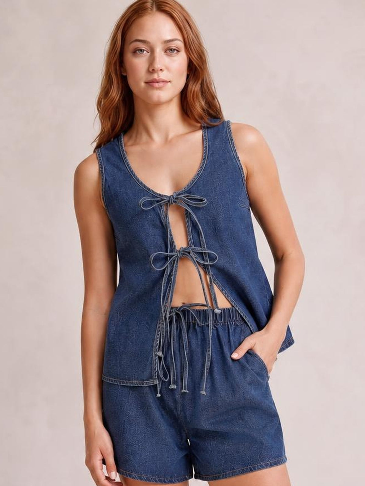 Model wearing a dark blue denim set with a sleeveless V-neck vest, multiple front bow ties, and relaxed shorts, posing with one hand in a pocket.