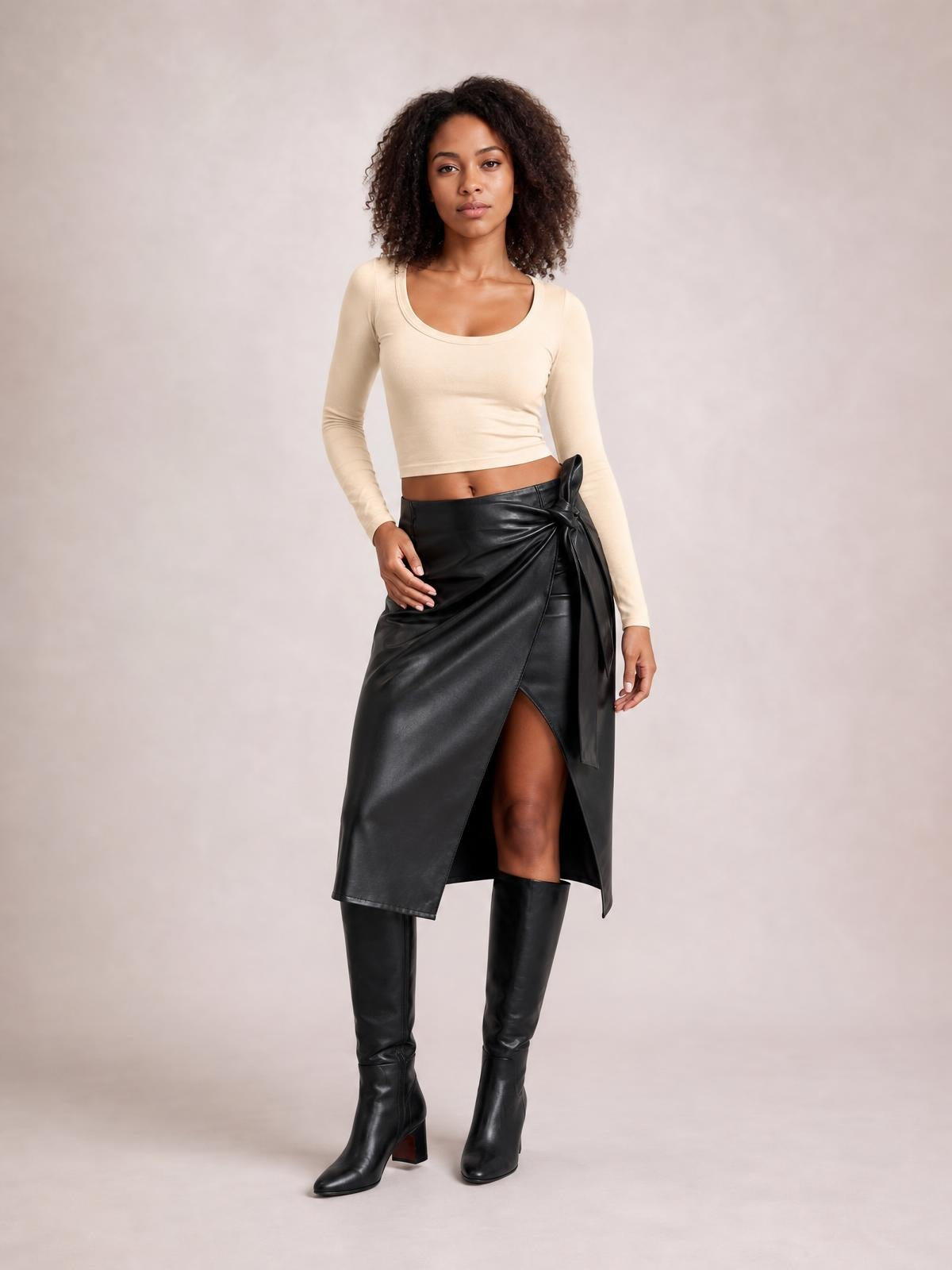 A model stands in a glossy black tied high waist skirt with a wrap silhouette, side bow detail, and front slit, paired with knee-high boots.