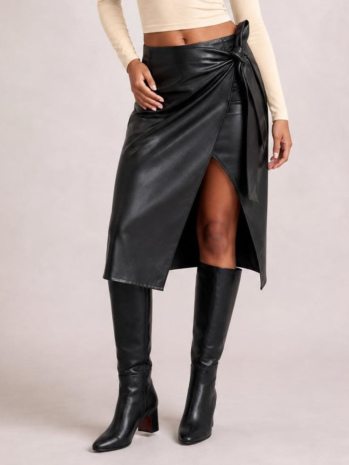 A model stands in a black tied high-waist skirt with a wrap-style front, draped construction, and thigh-high slit creating an asymmetric midi silhouette.