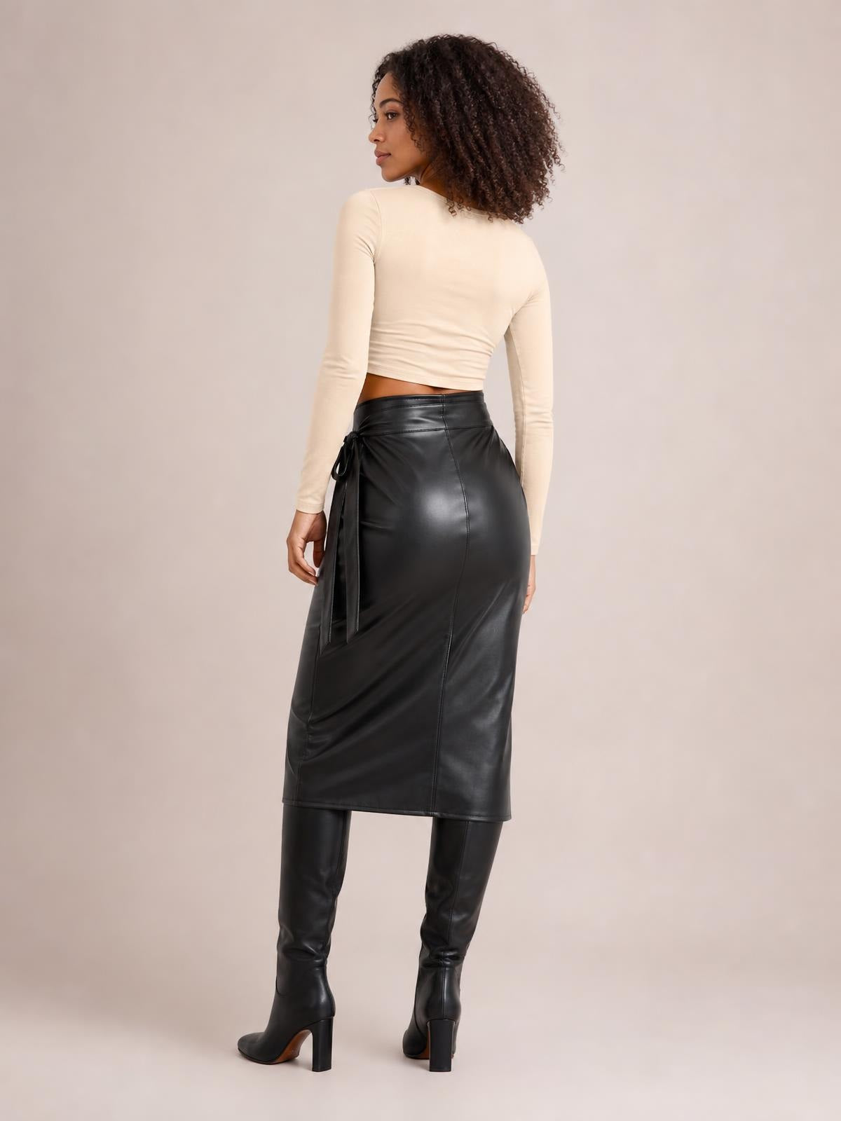 A model looks over her shoulder wearing a black high-waist skirt with a fitted midi silhouette, glossy finish, visible seams, and an adjustable side tie.