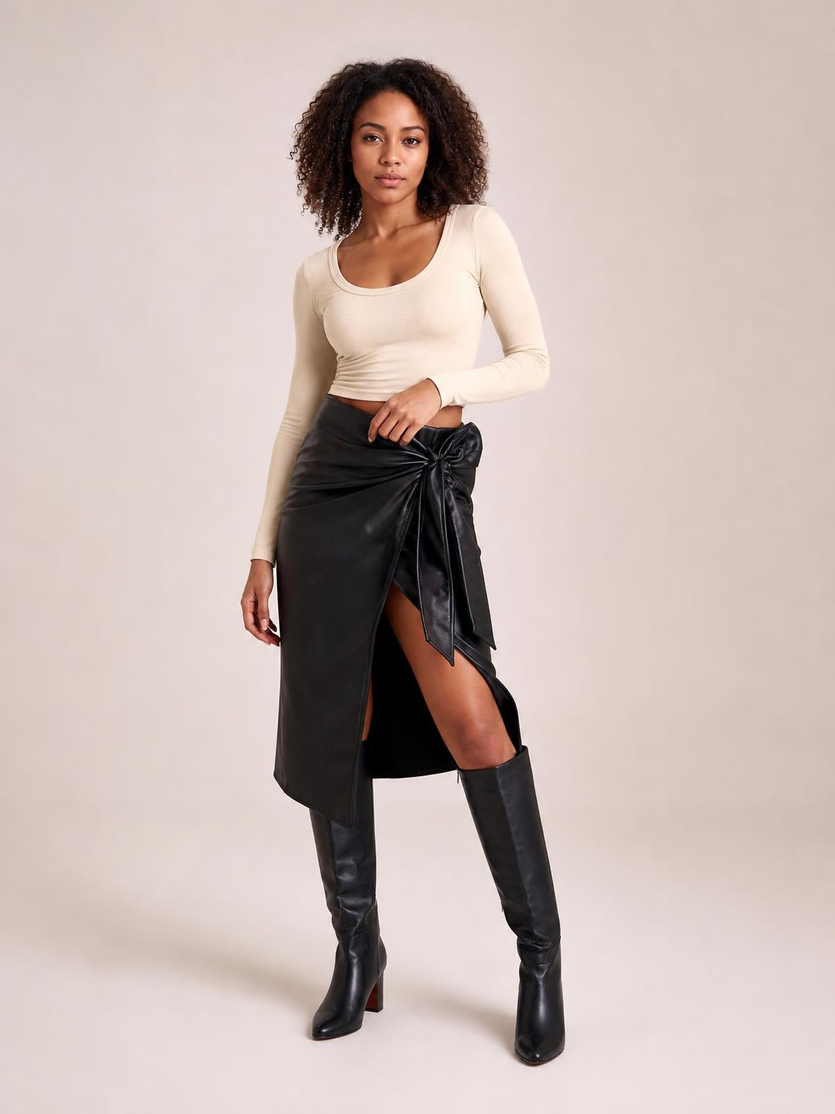 Model poses in a black high-waist skirt with an adjustable tied waist, sleek asymmetric wrap silhouette, and front slit revealing the leg.