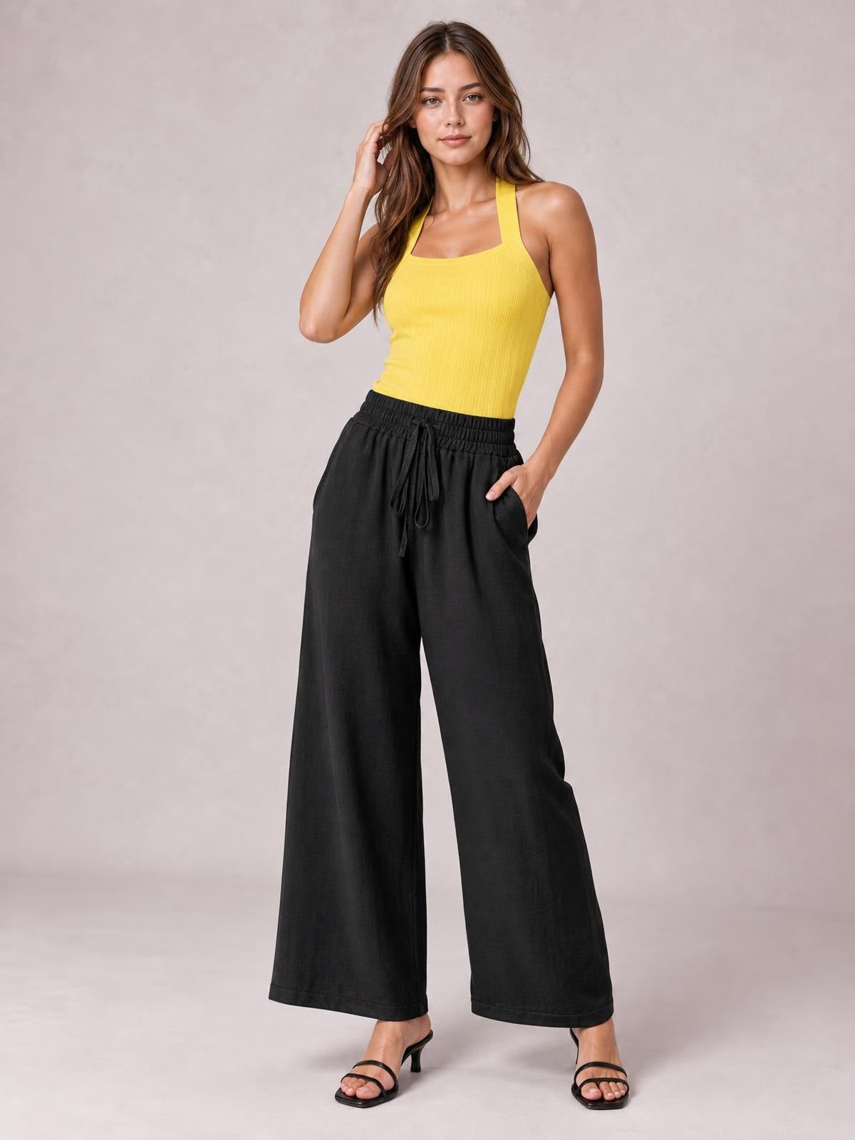 Model poses in black tied high waist wide leg pants with front pockets, an elastic waistband, relaxed flowing silhouette, and ankle-length hems.