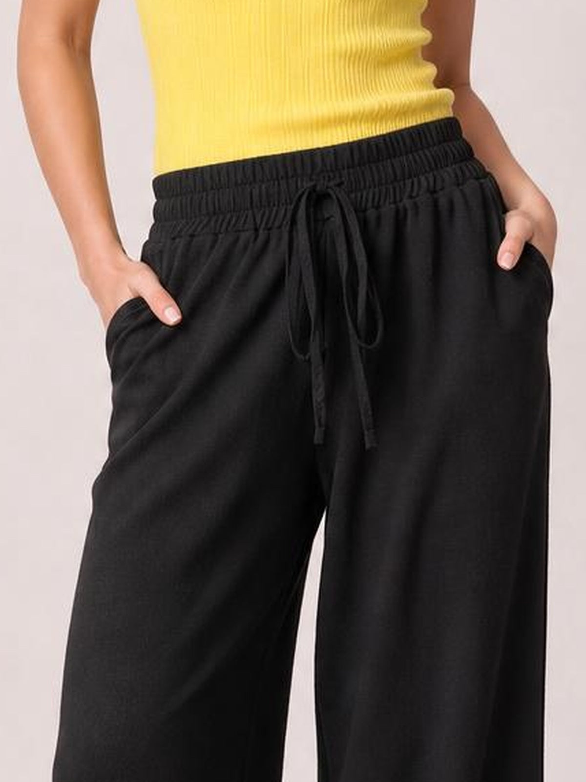 Model wearing black high-waist wide-leg pants with a gathered waistband, front tie, and side pockets, shown with hands resting inside the pockets.