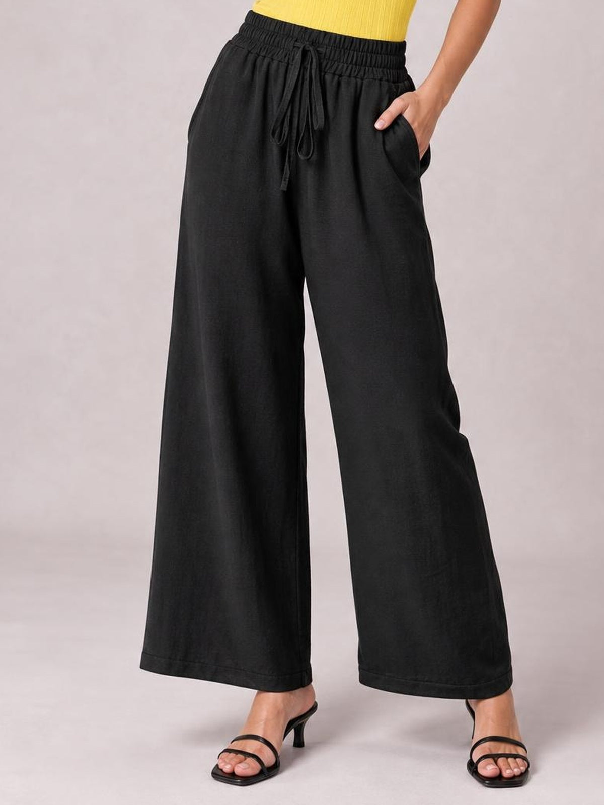 A model poses in black high-waist wide-leg pants with an elastic drawstring waist, side pockets, and a fluid full-length silhouette.
