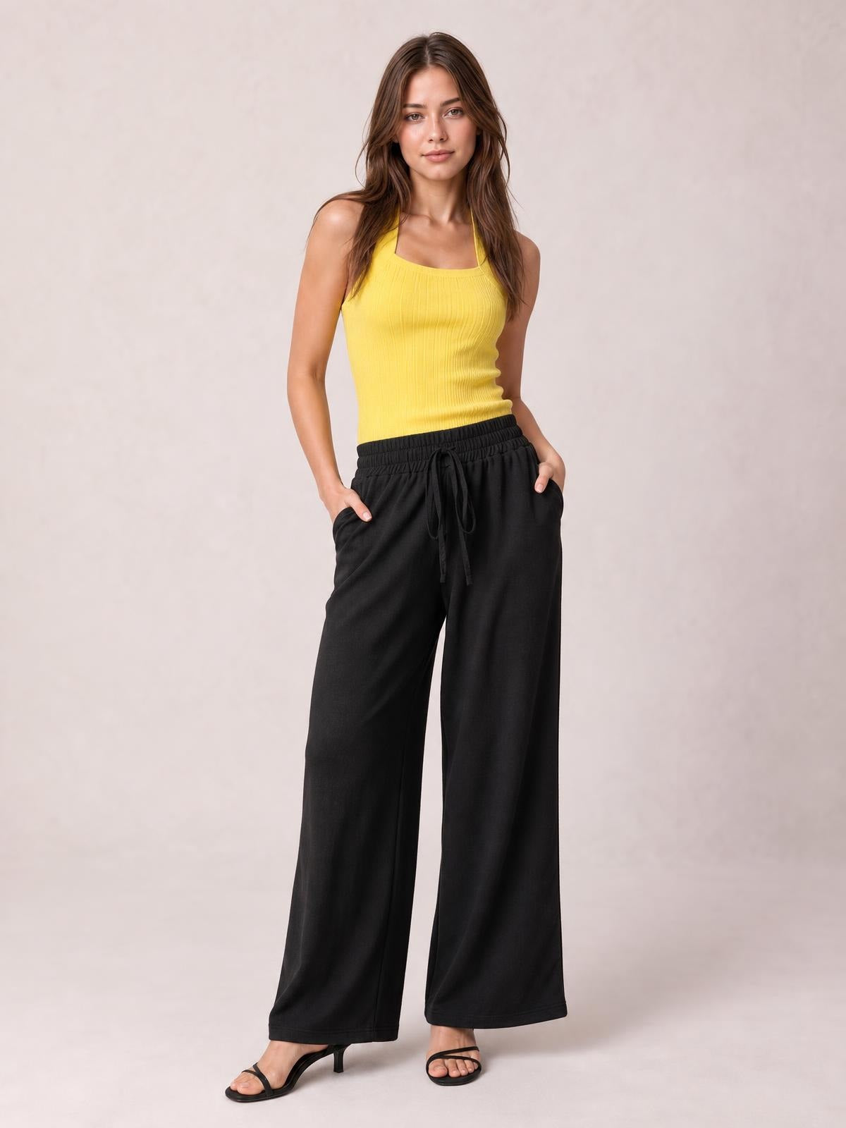 Model poses with hands in the pockets of black tied high-waist wide-leg pants, featuring a drawstring waist and relaxed floor-length silhouette.