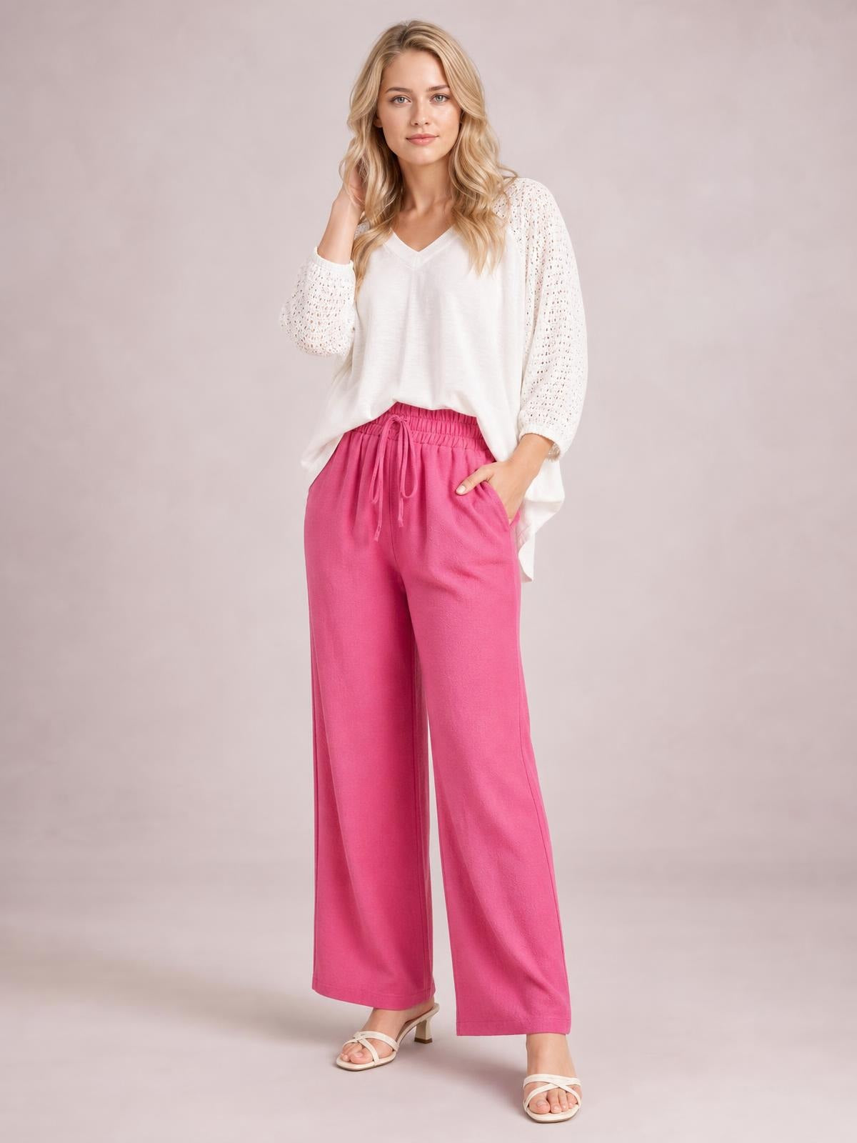 A model poses in bright pink high-waist wide-leg pants with a tied drawstring waistband and side pockets, standing with one hand tucked into a pocket.