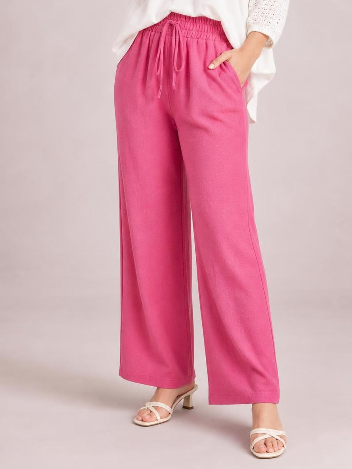 Model wearing bright pink high-waist wide-leg pants with a tied drawstring waist and side pockets, standing with one hand tucked into a pocket.