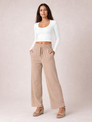 Model poses in beige tied high-waist wide-leg pants with an elastic waistband, drawstring, side pockets, and a relaxed full-length silhouette.