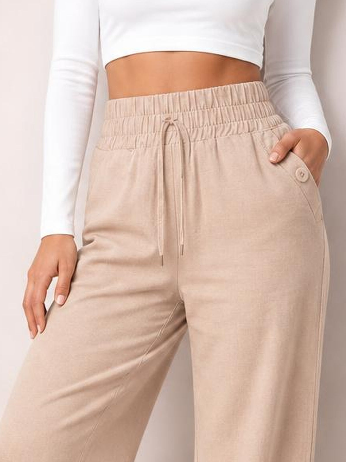 Model wearing beige high-waist wide-leg pants with a tied drawstring waist, decorative button pocket, and relaxed silhouette.