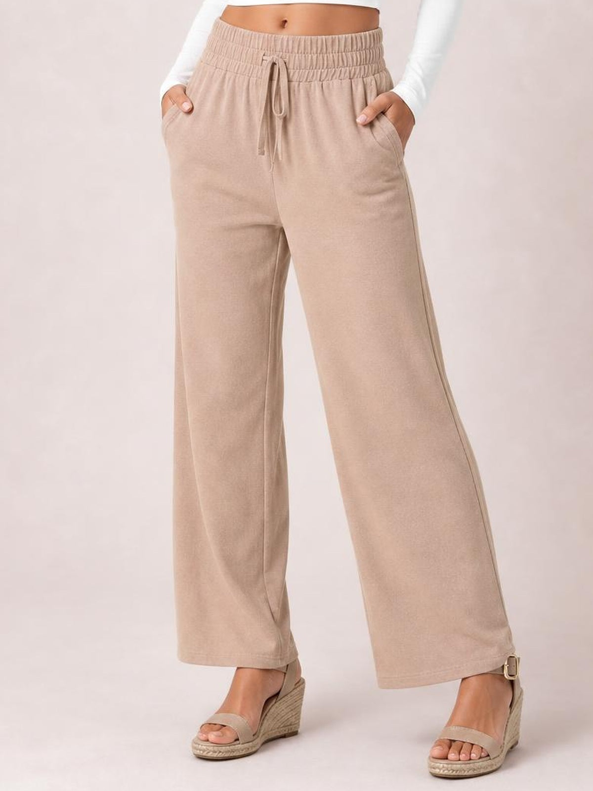 Model wearing beige high-waist wide-leg pants with an elastic waistband, tied drawstring, and side pockets, standing with hands tucked into the pockets.