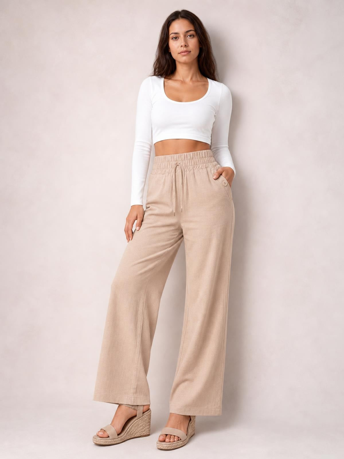 Model poses in beige tied high-waist wide-leg pants with an elastic waistband, drawstring, side pockets, and decorative buttons.