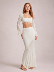Model poses in a white openwork cover-up set with a square-neck tied long-sleeve crop top and high-waisted fitted maxi skirt.