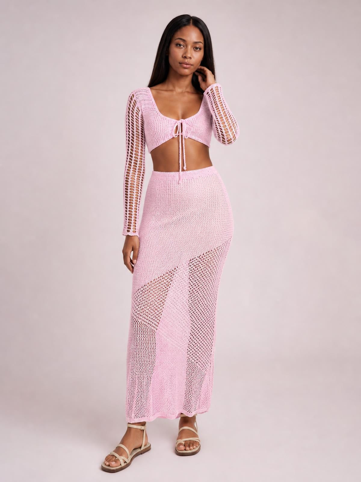 Model wearing a pink tied long sleeve top and matching openwork maxi skirt cover-up set, with a cropped silhouette and relaxed standing pose.