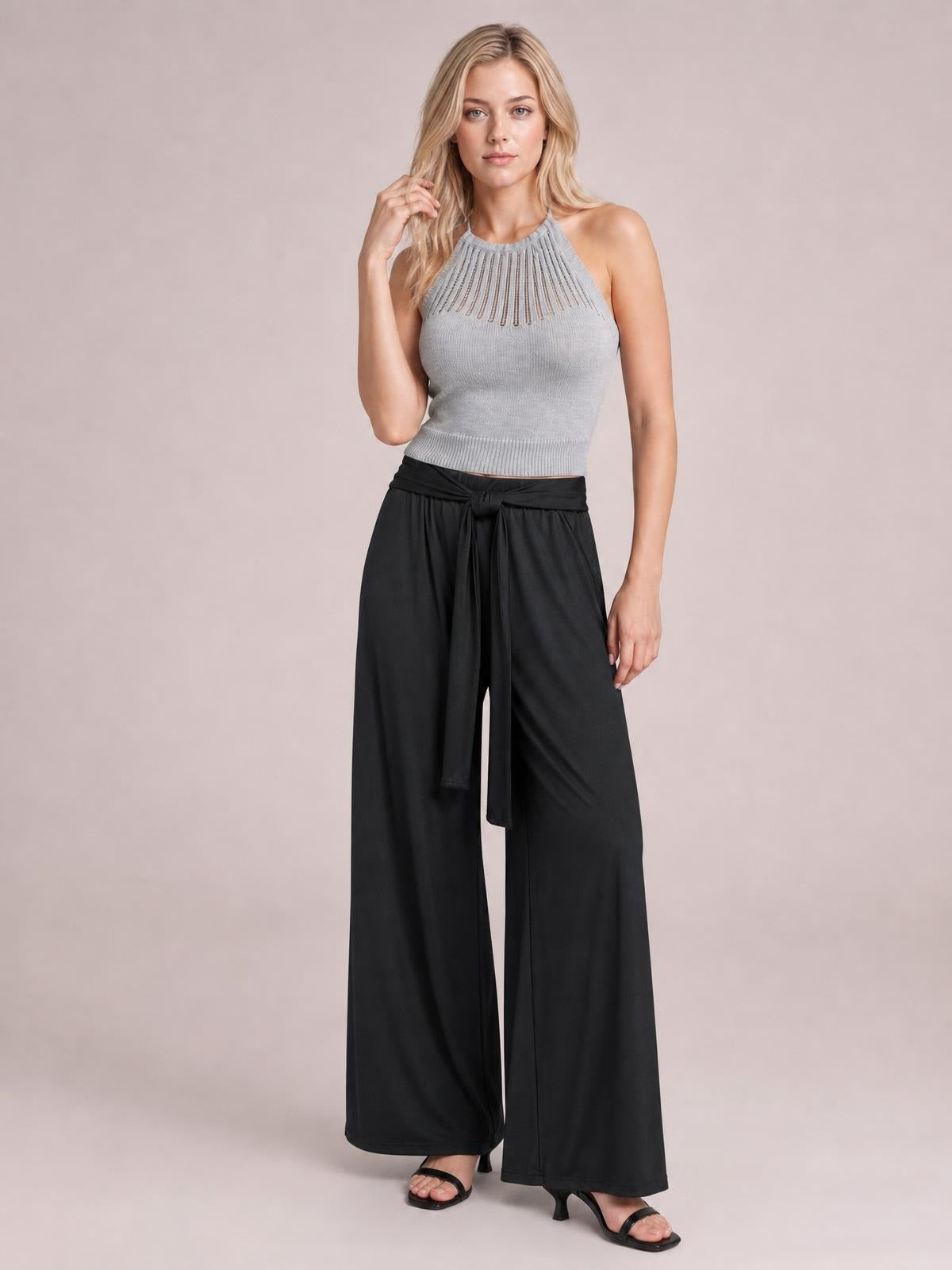 A standing model wears black tied pocketed wide-leg pants with a flowing full-length silhouette and relaxed drape, posing with one hand lifted to her hair.