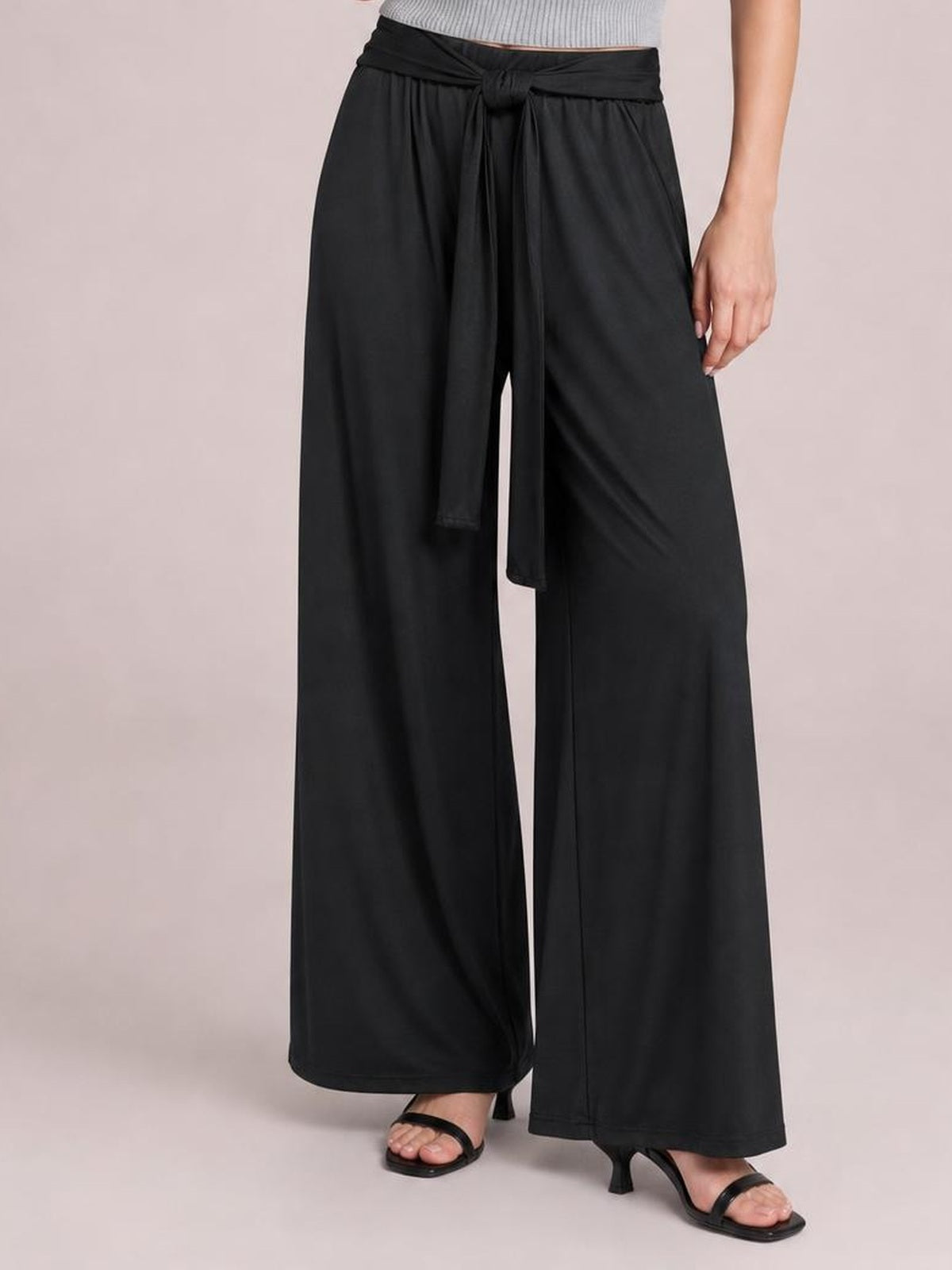 A standing model wears black opaque wide-leg pants with a tied waist, long sash ends, and practical side pockets in a relaxed pose.