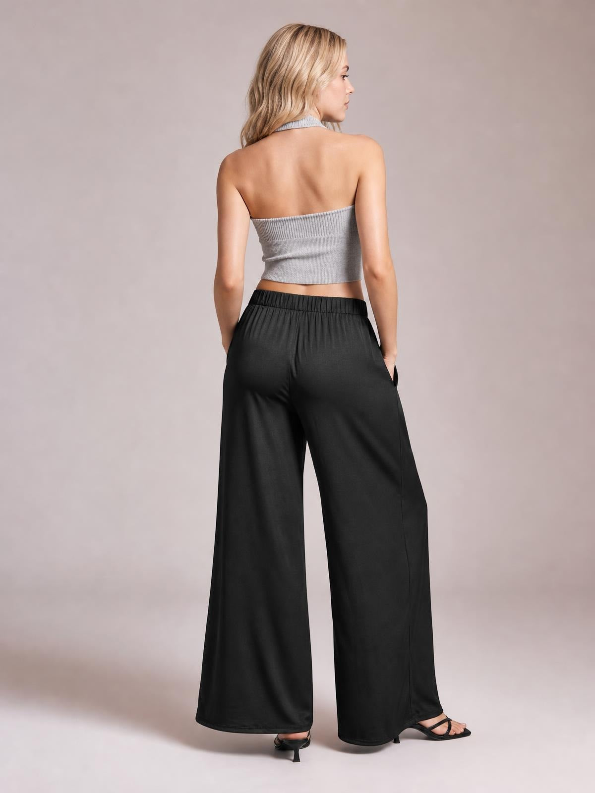 A model poses from the back in black tied pocketed wide-leg pants with an elastic waistband, flowing full-length silhouette, and hands in the pockets.