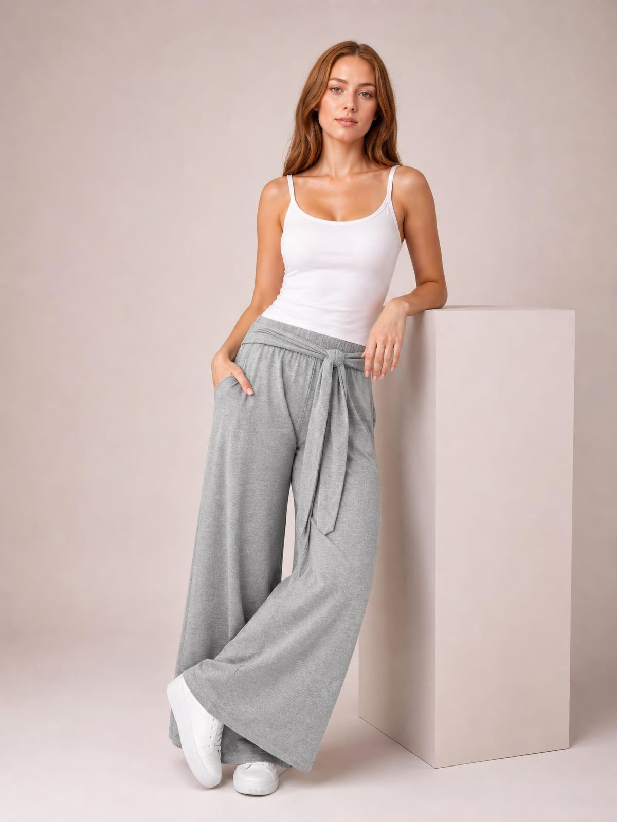 Model poses in gray tied pocketed wide leg pants with an elastic waist, flowing full-length silhouette, and hands tucked into the side pockets.